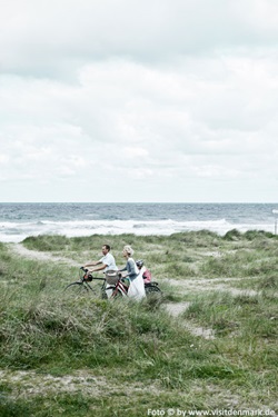 Eine kleine Familie schiebt ihre Räder durch die Dünen Dänemarks, das Kind sitzt auf dem Gepäckträger eines Fahrrads, im Hintergrund ist das Meer mit brechenden Wellen zu sehen