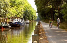 Zwei Radfahrer radeln auf einem Radweg neben einem Kanal in der Provinz Limburg entlang, in dem einige Schiffe und Boote angelegt sind