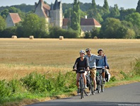 Drei RadlerInnen fahren auf einem Radweg an einem gemähten Getreidefeld bei Courceraulp vorbei. Im Hintergrund erhebt sich das Herrenhaus "Manoir de Veuve".