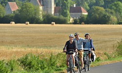 Drei RadlerInnen fahren auf einem Radweg an einem gemähten Getreidefeld bei Courceraulp vorbei. Im Hintergrund erhebt sich das Herrenhaus "Manoir de Veuve".