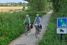 Ein Radlerpärchen fährt auf einem weiß geschotterten Radweg durch ein kleines Wäldchen bei Conceraulp. Im Hintergrund ist das Herrenhaus "Manoir de Veuve" zu erkennen.