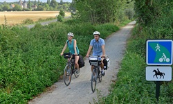 Ein Radlerpärchen fährt auf einem weiß geschotterten Radweg durch ein kleines Wäldchen bei Conceraulp. Im Hintergrund ist das Herrenhaus "Manoir de Veuve" zu erkennen.