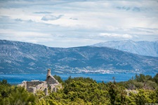 Die von dichtem Gebüsch eingerahmte Ruine eines verlassenen Hauses auf der Insel Hvar erhebt sich vor dem tiefblauen Meer und einer dahinter aufragenden Gebirgskette.