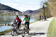 Insgesamt 6 Radler, davon eine Familie mit Kind, radeln auf dem Donauradweg am Flussufer entlang.