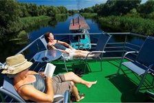 Zwei Passagiere genießen die Sonne auf dem Sonnendeck - ein liest ein Buch, währen das Floßhotel vom Schiff über die Donau gezogen wird