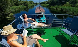 Zwei Passagiere genießen die Sonne auf dem Sonnendeck - ein liest ein Buch, währen das Floßhotel vom Schiff über die Donau gezogen wird