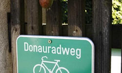 Grünes Schild des Donauradwegs: Schriftzug Donauradweg, darunter ein Fahrrad, links unter dem Fahrrad die Nr. 6 in einem gelben Kasten und daneben ein weißer Pfeil, der nach rechts zeigt
