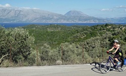 Eine Radlerin fährt auf einem asphaltierten Radweg über eine der Ionischen Inseln. Am Wegesrand wächst Gebüsch, im Hintergrund ist das tiefblaue Meer zu erkennen.