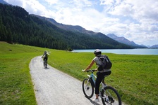 Zwei Radler fahrne auf einem weiß gekiesten Schotterweg am Silvaplanersee im Oberengadin entlang.