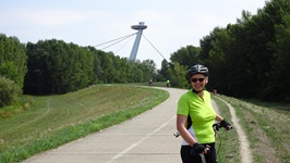 Eine Radfahrerin steht auf dem Donau-Radweg bei Bratislava, blickt nach hinten in die Kamera und im Hintergrund ist der bekannte UFO Aussichtsturm von Bratislava zu sehen ist