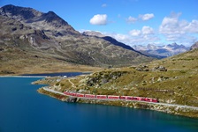 Ein roter Regionalzug fährt am Ufer des Lago Bianco entlang; im Hintergrund erhebt sich das Bernina-Massiv.