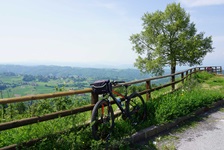 Ein abgestelltes Fahrrad lehnt in Ligurien an einem Koppelgatter. Im Hintergrund sind saftig grüne Wiesen und eine im Dunst liegende Bergkette zu erkennen.