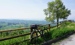Ein abgestelltes Fahrrad lehnt in Ligurien an einem Koppelgatter. Im Hintergrund sind saftig grüne Wiesen und eine im Dunst liegende Bergkette zu erkennen.