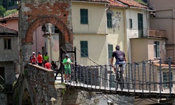 Ein Radfahrer radelt über eine kleine Hängebrücke auf das mittelalterliche Stadttor von Millesimo zu.