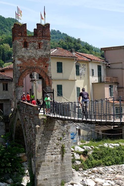 Ein Radfahrer radelt über eine kleine Hängebrücke auf das mittelalterliche Stadttor von Millesimo zu.