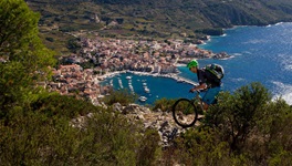Ein Mountainbiker auf der Insel Vis fährt über einen sehr steinigen Naturweg hinunter zum einladend blau schimmernden Meer und einer kleinen Siedlung.