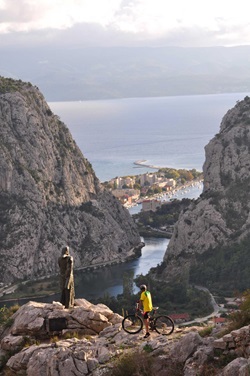 Ein Mountainbiker steht auf einem Aussichtsfelsen mit Statue in Omis und blickt auf den Canyon und die Mündung der Cetina in das Adriatische Meer in Süddalmatien