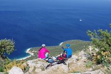 Zwei Mountainbiker sitzen in Süddalmatien auf einer Klippe und schauen hinunter auf eine Bucht mit tiefblauem Wasser, in der ein kleines Schiff ankert.