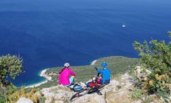 Zwei Mountainbiker sitzen in Süddalmatien auf einer Klippe und schauen hinunter auf eine Bucht mit tiefblauem Wasser, in der ein kleines Schiff ankert.