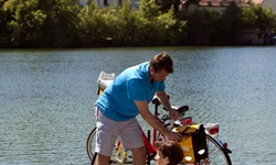 Zwei Radfahrer gönnen sich eine Pause auf einem Holzsteg - die Frau sitzt auf dem Steg und zieht ihren Fuß aus dem Wasser, währen der Mann die Gepäcktasche am Rad aufmacht - im Hintergrund ist das Kloster Seeon mit seinen zwei Zwiebeltürmen zu sehen