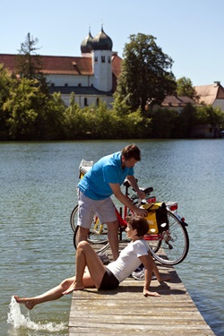 Zwei Radfahrer gönnen sich eine Pause auf einem Holzsteg - die Frau sitzt auf dem Steg und zieht ihren Fuß aus dem Wasser, währen der Mann die Gepäcktasche am Rad aufmacht - im Hintergrund ist das Kloster Seeon mit seinen zwei Zwiebeltürmen zu sehen