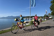 Radfahrer radeln an der Promenade des Chiemsees entlang, während im Hintergrund die Alpen zu sehen sind