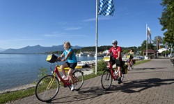 Radfahrer radeln an der Promenade des Chiemsees entlang, während im Hintergrund die Alpen zu sehen sind