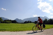 Ein Fahrradfahrer radelt auf einem geschotterten Weg an Wiesen und Wälder vorbei und hat einen traumhaften Blick auf die Alpen