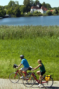 Zwei Radfahrer radeln am Klostersee in Seeon vorbei - im Hintergrund ist die Kirche zu sehen