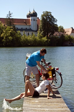 Zwei Radfahrer gönnen sich eine Pause am Klostersee - im Hintergrund ist das ehemalige Kloster Seeon mit seinen zwei Zwiebeltürmen zu sehen