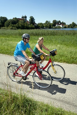 Radfahrer radeln auf einem asphaltierten Radweg am Klostersee in Seeon vorbei - im Hintergrund ist das ehemalige Kloster Seeon mit seinen beiden Zwiebeltürmen und die Kirche zu erkennen