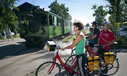 Vier Radfahrer warten bis die grüne Chiemsee Bahn den unbeschrankten Bahnübergang passiert hat