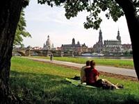 Ein Pärchen sitzt auf einer Decke unter einem Baum am Elbufer und genießt den Blick auf die Skyline von Dresden.