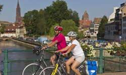 Zwei Radler - ein Mann und eine Frau - fahren über eine mit Blumen geschmückte Brücke in Straßburg. Im Bildhintergrund ist der Turm des Münsters zu erkennen.