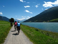 Eine Radlergruppe fährt auf dem parallel zur Etsch verlaufenden Etschtal-Radweg auf die schneebedeckten Gipfel der Dolomiten zu.