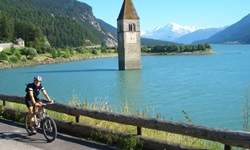Ein Radfahrer fährt auf dem Etschtal-Radweg am Reschensee und dem versunkenen Kirchturm von Graun vorbei.