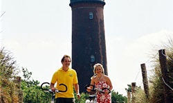 Zwei Radler schieben ihre Räder - im Hintergrund ist ein Leuchtturm der Ostsee zu sehen