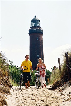 Zwei Radler schieben ihre Räder - im Hintergrund ist ein Leuchtturm der Ostsee zu sehen