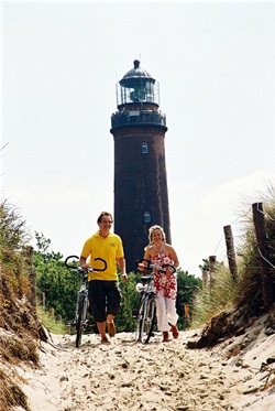 Zwei Radler schieben ihre Räder auf einem Sandweg, hinter ihnen ist ein Leuchtturm zu sehen