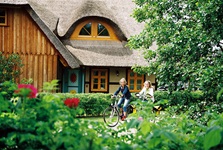 Zwei Radfahrerinnen radeln an einem typischen Haus an der Ostsee mit Reetdach vorbei