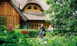 Zwei Radfahrerinnen radeln an einem typischen Haus an der Ostsee mit Reetdach vorbei