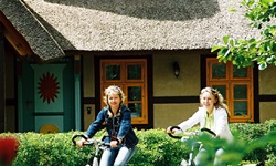 Zwei Radlerinnen fahren an einem Haus mit Reetdach vorbei