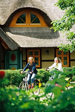 Zwei Radlerinnen fahren an einem Ostsee-Haus mit Reetdach vorbei