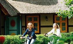 Zwei Fahrradfahrerinnen radeln an eine Haus mit Reetdach vorbei