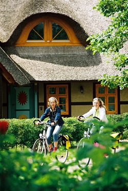 Zwei Fahrradfahrerinnen radeln an eine Haus mit Reetdach vorbei