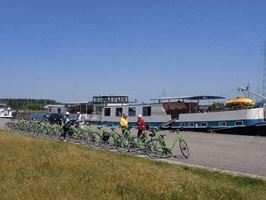 Für Gäste vorbereitete grüne Fahrräder stehen auf einem Radweg bereit, währen die ehemalige MS Fiep an einem Ufer liegt