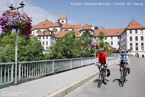 Ein Radler-Paar fährt auf der Theresienbrücke in Füssen auf der Via Claudia Augusta - im Hintergrund ist das Kloster St. Mang