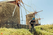 Ein Mountainbiker nimmt auf Patmos vor der traumhaften Kulisse von drei Windmühlen eine von blühenden Wiesen gesäumte Downhill-Strecke in Angriff.