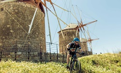 Ein Mountainbiker nimmt auf Patmos vor der traumhaften Kulisse von drei Windmühlen eine von blühenden Wiesen gesäumte Downhill-Strecke in Angriff.