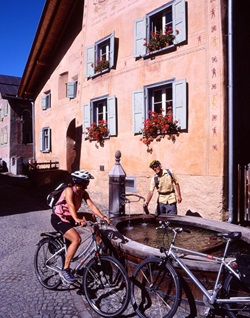 Ein Radlerpärchen macht an einem Brunnen in einem Engadiner Dorf Pause.